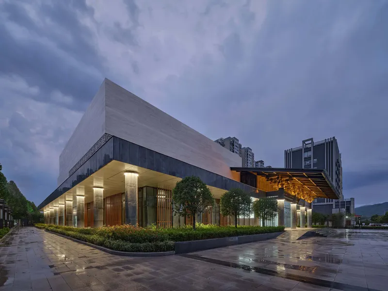 Corner view of the illuminated entrance colonnade with timber screens and planted beds at dusk after rain