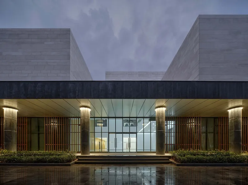 Entrance canopy with glass doors framed by lit timber screens and planted beds beneath a volumetric ceiling at twilight