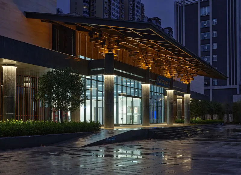 Night view of the illuminated glass entrance beneath a sculptural timber soffit canopy with wet paving reflecting light