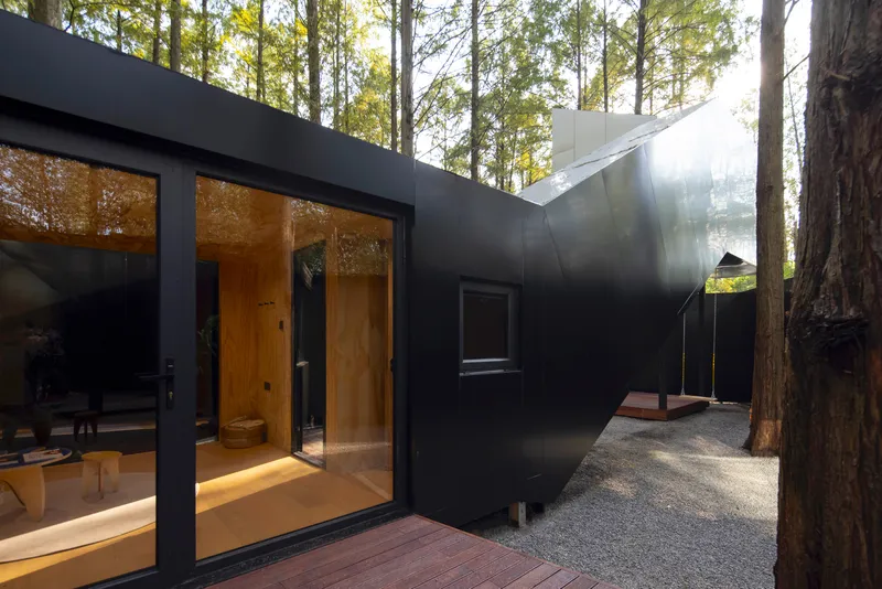 Black metal-clad exterior with timber deck and full-height glazing nestled among tall forest trees