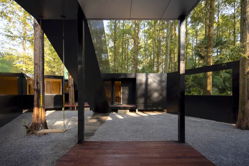 View from the timber deck through the courtyard gate toward the black steel volumes and preserved eucalyptus trunks