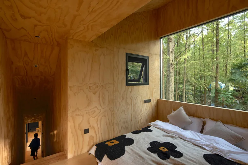 Plywood-lined bedroom with a figure descending the stairs and a ribbon window framing the forest beyond