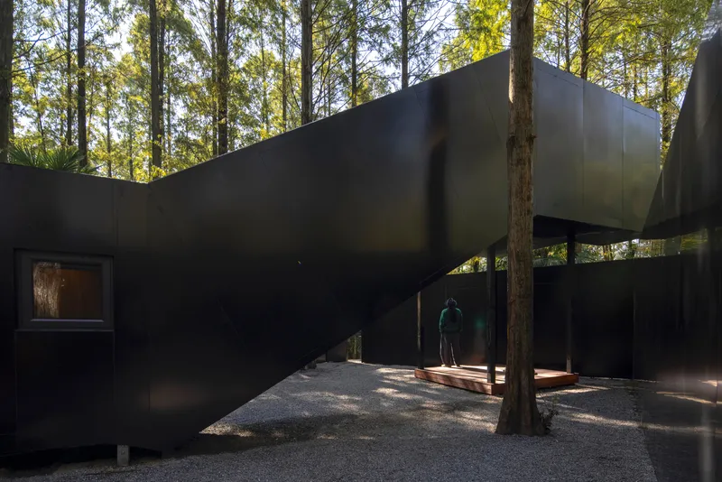 Covered walkway with undulating black metal walls and a figure standing beneath filtered afternoon light