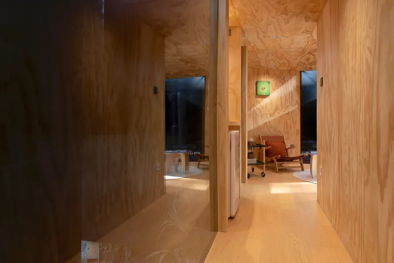 Plywood corridor with pivoting door leading to living area with green clock on wall