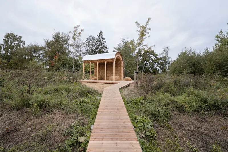 Timber pavilion with arched straw bale walls reached by a wooden boardwalk through planted landscape