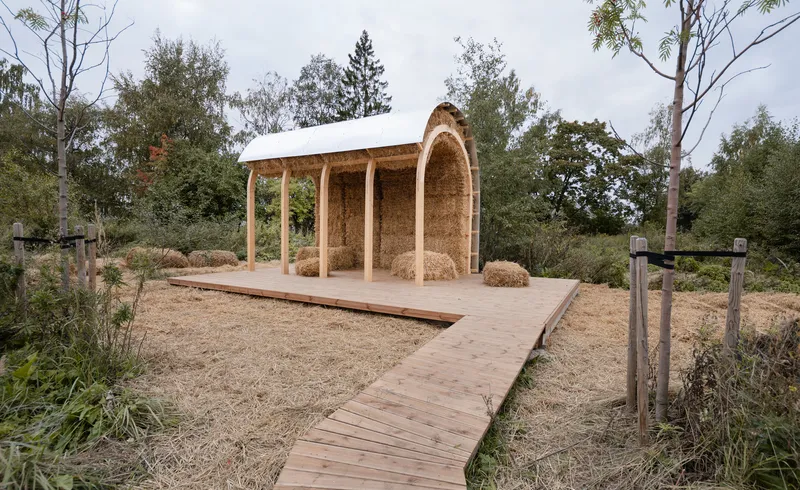 Open pavilion with timber columns and straw bale walls beneath a flat roof on a gravel clearing