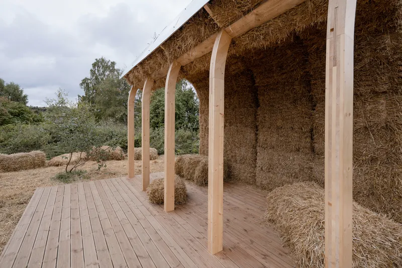 Timber deck and columns supporting a straw bale roof with trees visible in the background