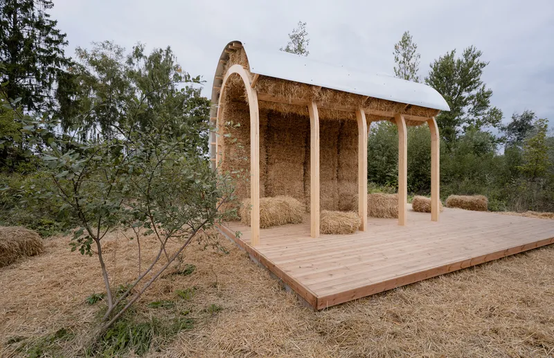 Arched straw bale pavilion with timber deck and white tensile roof under an overcast sky