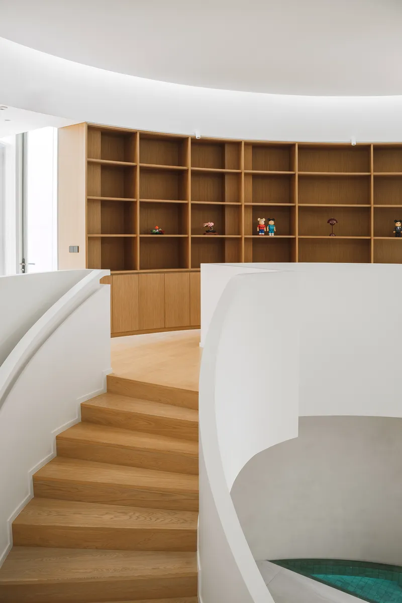 Curved timber staircase ascending toward a floor-to-ceiling oak shelving system with scattered figurines