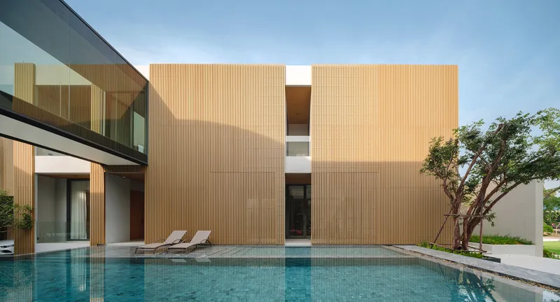 Poolside view of the timber-clad volume with glazed bridge and stucco wall beside a mature tree