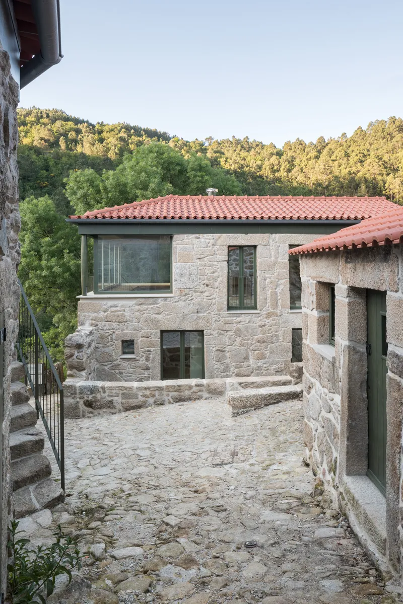 Narrow cobblestone lane descending between stone volumes with glazed openings and balconies overlooking the valley