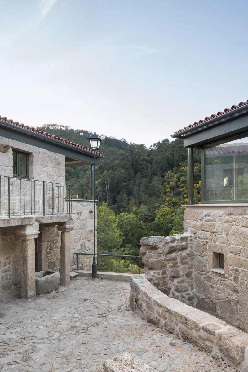 Stone courtyard terrace with historic column fragments and contemporary glazing frames opening to the wooded hillside at dusk