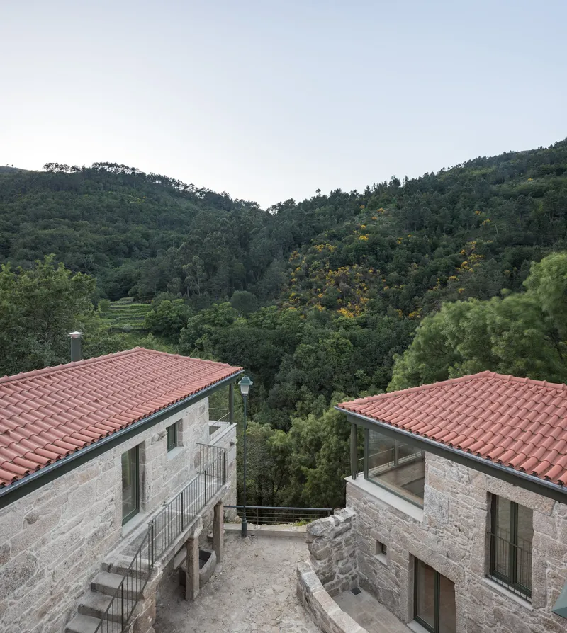 Courtyard between two stone volumes with terracotta roofs opening toward the valley beyond at dusk