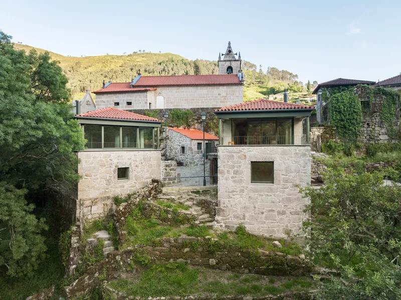 Exterior view of stone volumes with terracotta roofs and glazed openings set among vegetation and rocky terrain