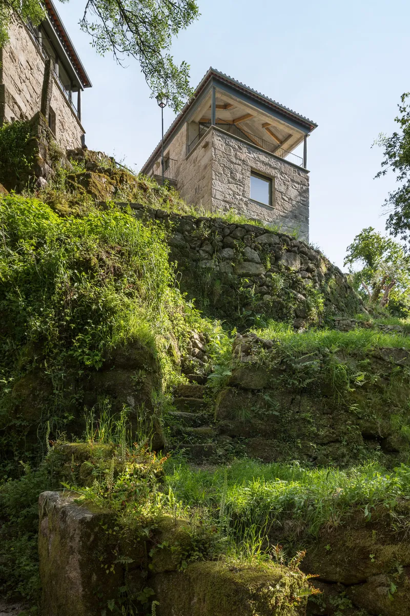 Stone tower with exposed timber roof structure rising above a moss-covered hillside garden