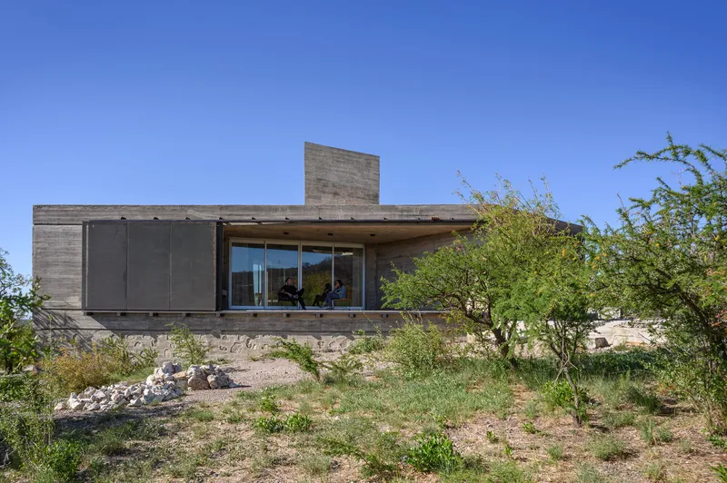 Board-formed concrete house with horizontal facade and metal screens on a sloped grassy site with fruit trees