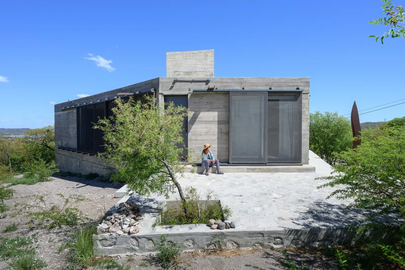 Entrance facade with board-formed concrete walls, mesh screens, and two figures at the entry under blue sky
