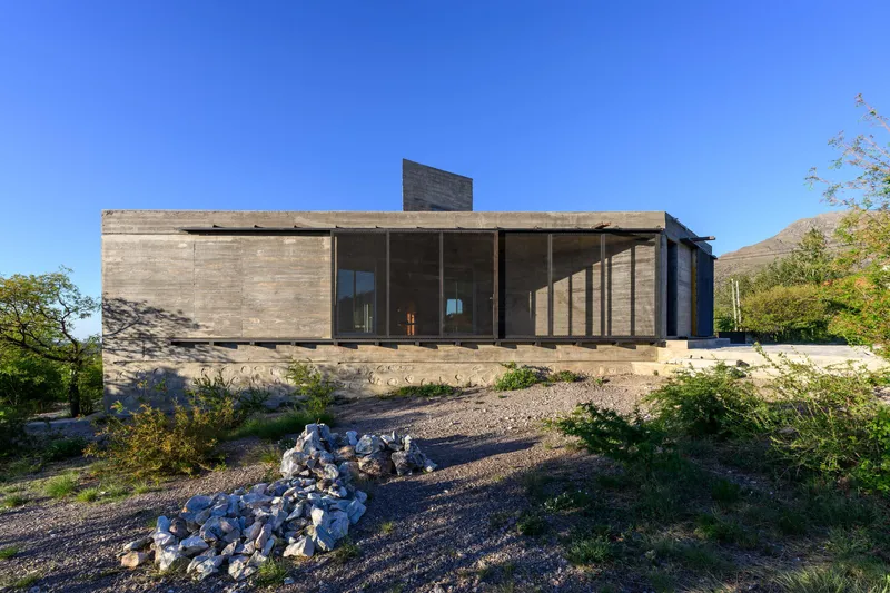 Boardform concrete facade with cantilevered upper volume overlooking a gravel landscape with scattered white rocks