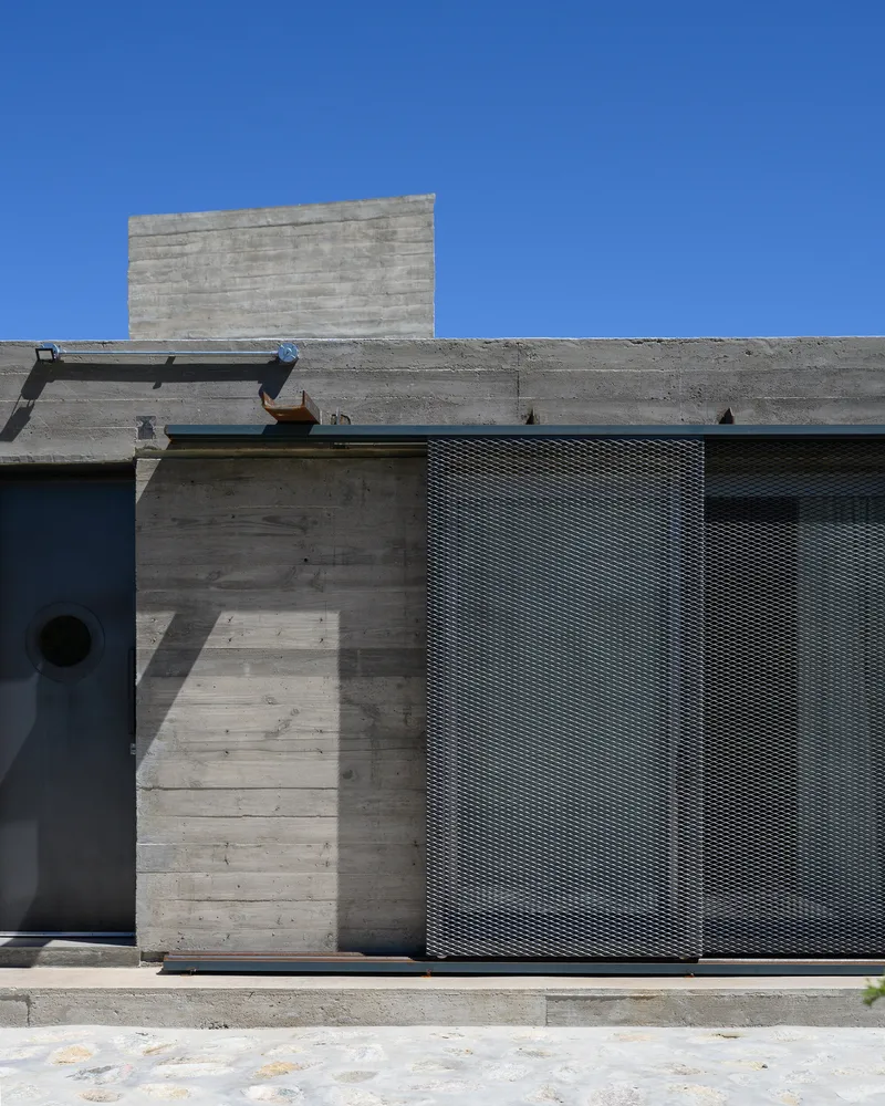 Facade detail showing boardform concrete walls with metal mesh screen panel under a clear blue sky