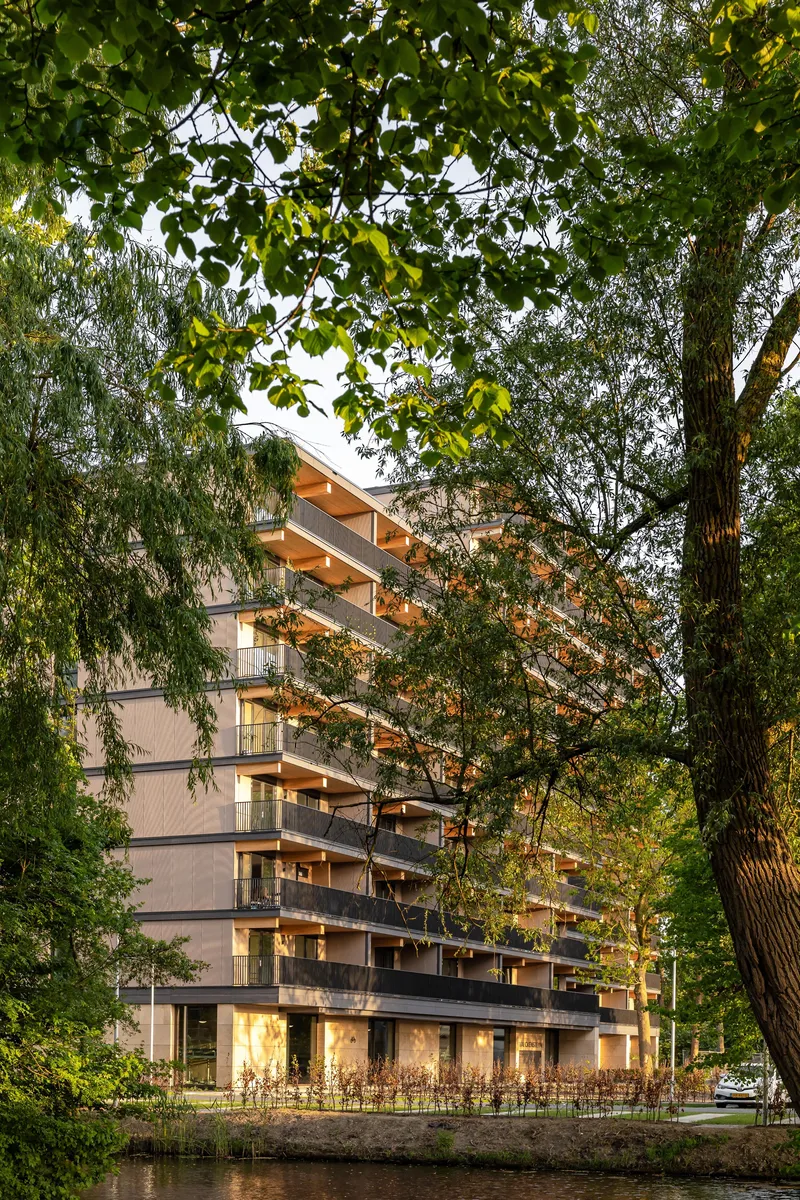 Facade with cantilevered timber beams viewed through willow and deciduous tree branches at twilight