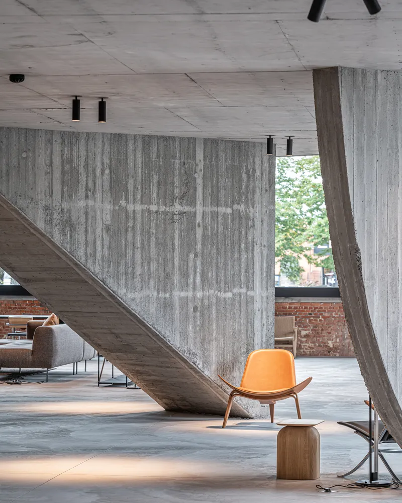 Board-formed concrete stair underside with orange upholstered chair and angled concrete column