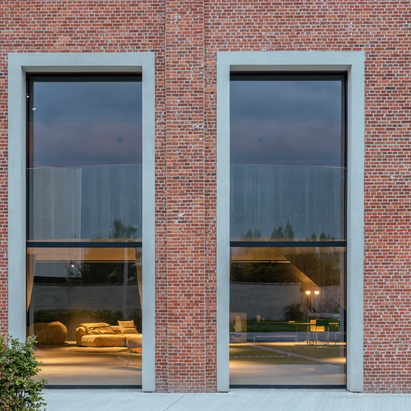 Two tall glazed openings framed in pale plaster reveal interior lounge furniture at dusk