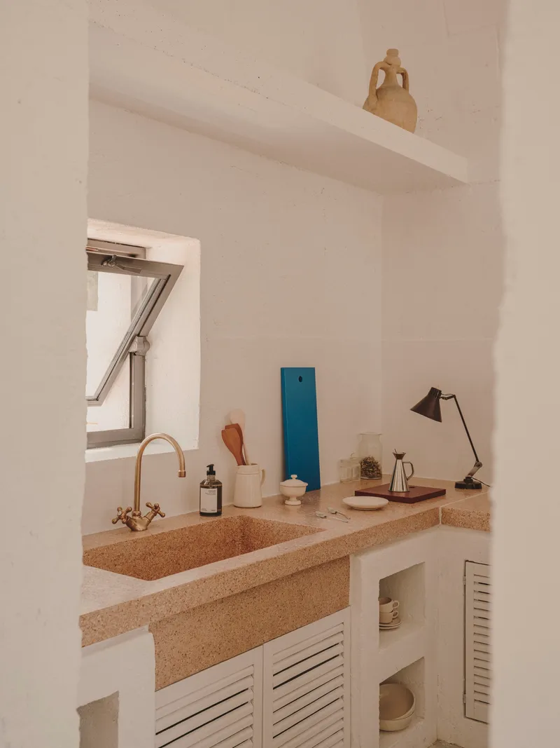 Kitchen counter with terrazzo surface, brass faucet and louvered cabinet doors below skylight