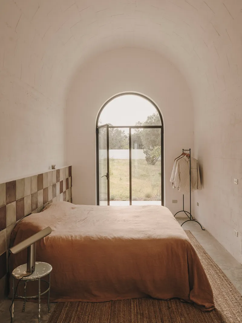 Bedroom with patchwork timber headboard and arched steel door framing view of olive grove