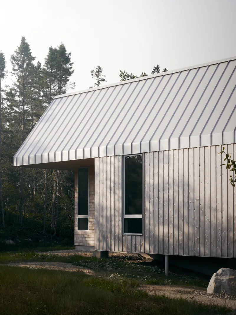 Gable-roofed volume with vertical timber siding elevated on concrete piers among evergreens