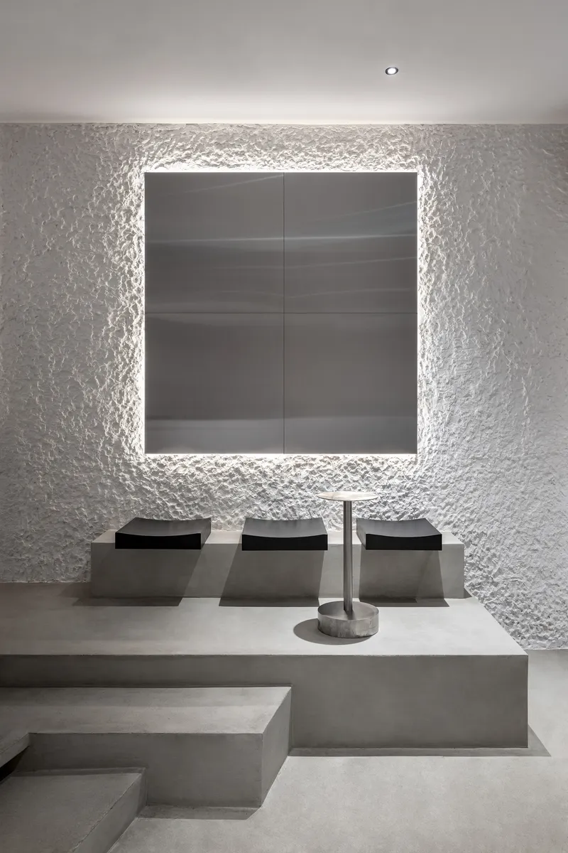 Stepped concrete seating platform facing backlit dark panel framed by rough white plastered walls