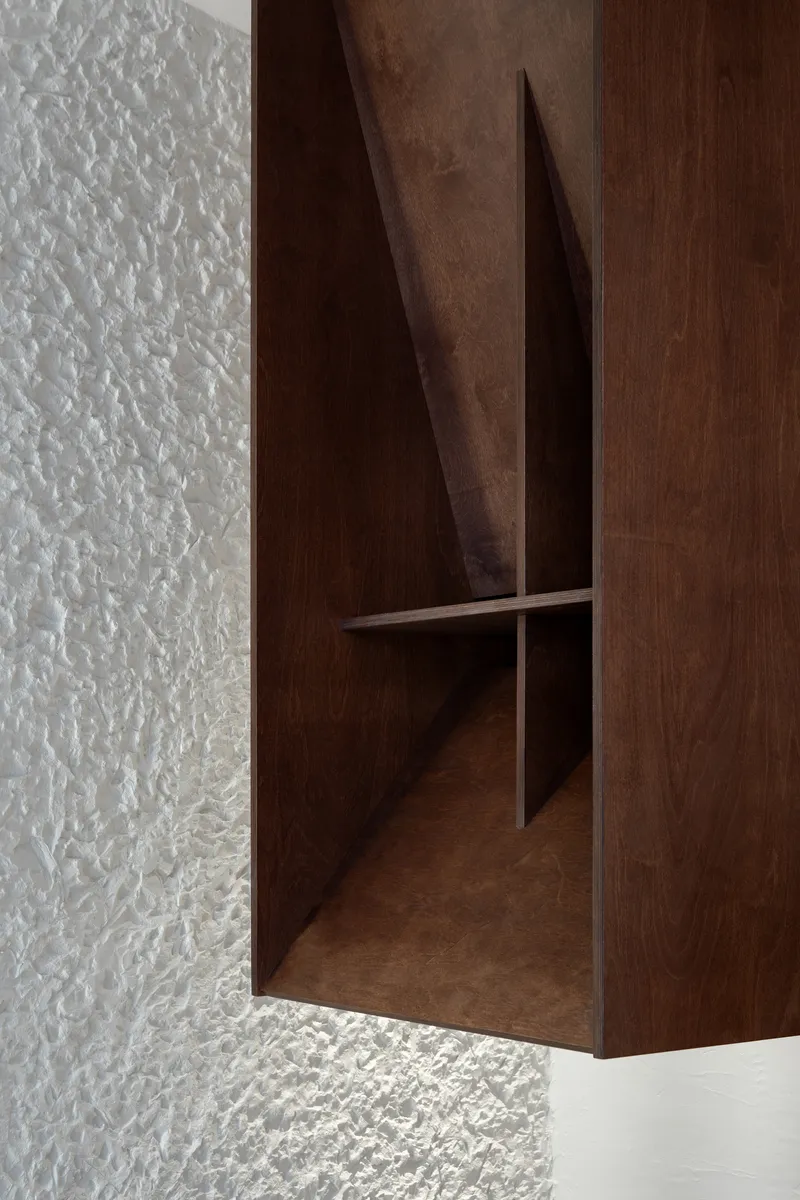 Triangular opening in a suspended walnut cabinet revealing interior shelving against textured plaster