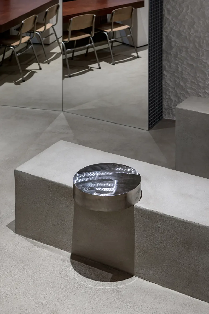 Polished metal cylindrical stool on a raised concrete platform beneath mirrored walls and folding chairs