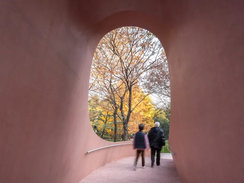Interior tunnel view through arched pink concrete passage framing two visitors and autumn foliage beyond