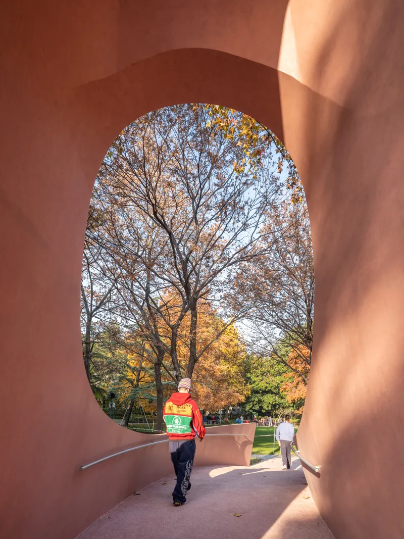 Visitor in red jacket walking through arched pink passage framing autumn landscape and park visitors