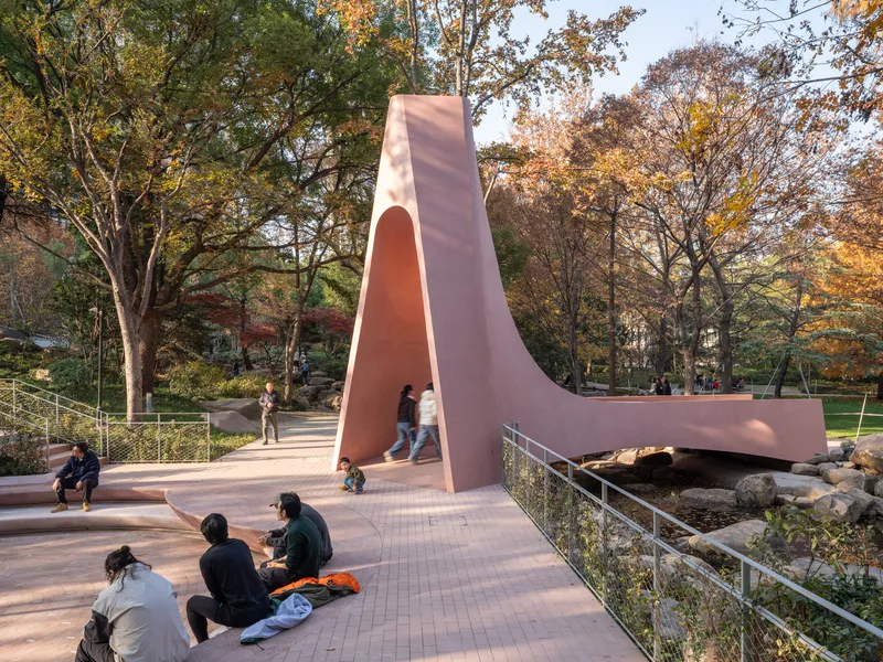 Sculptural pavilion with arched opening spanning a park pathway under dappled autumn sunlight