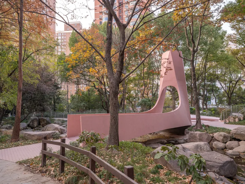 Pink sculptural pavilion spanning boulders and a dry streambed among autumn trees in a park