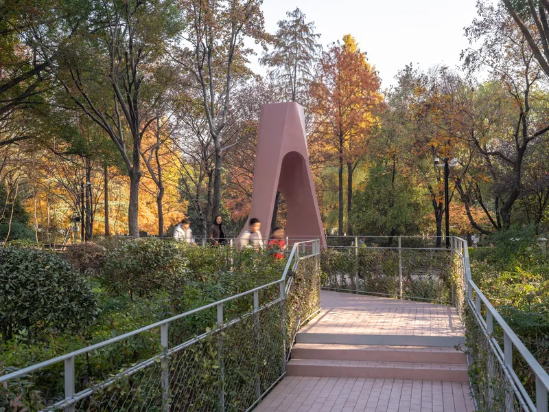 Mesh-railed pedestrian bridge leading to the pink arch structure at dusk with backlit trees
