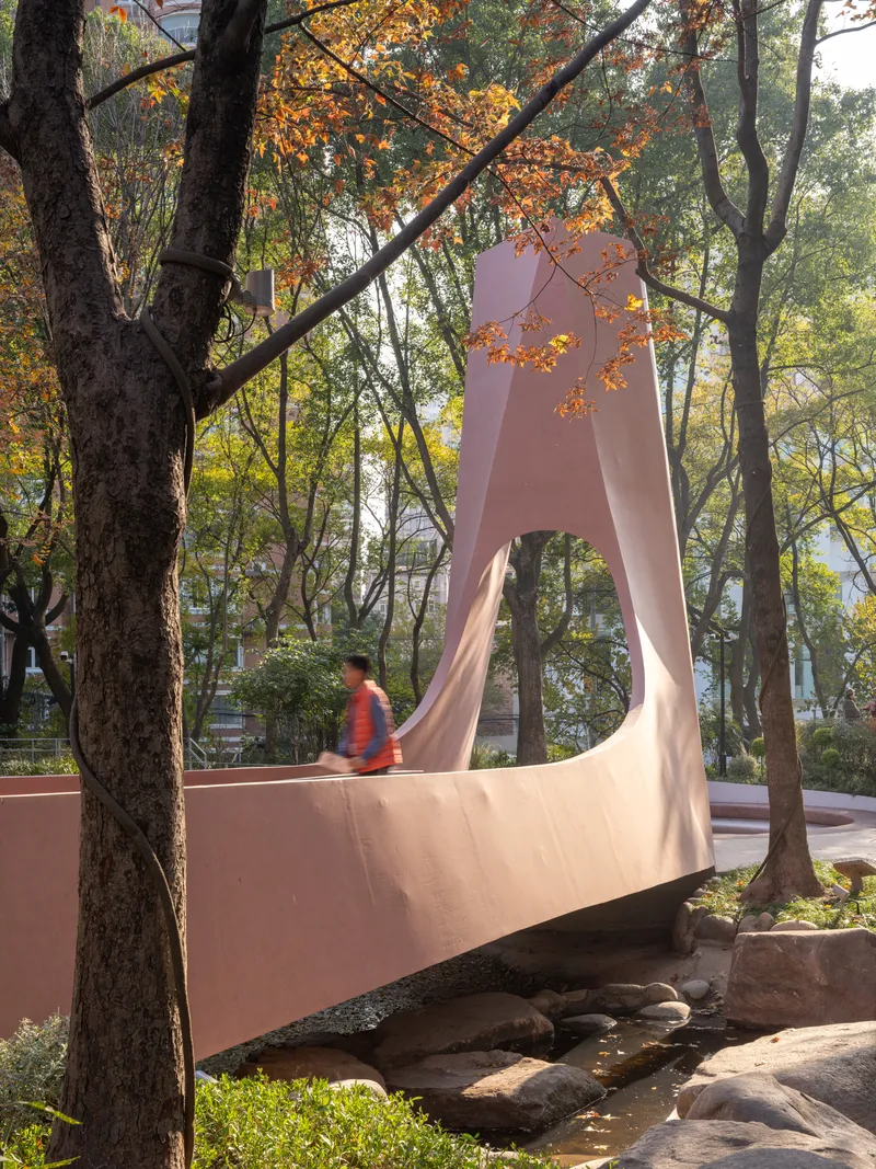 Pink sculptural footbridge spanning a rocky stream bed with a person crossing among autumn trees