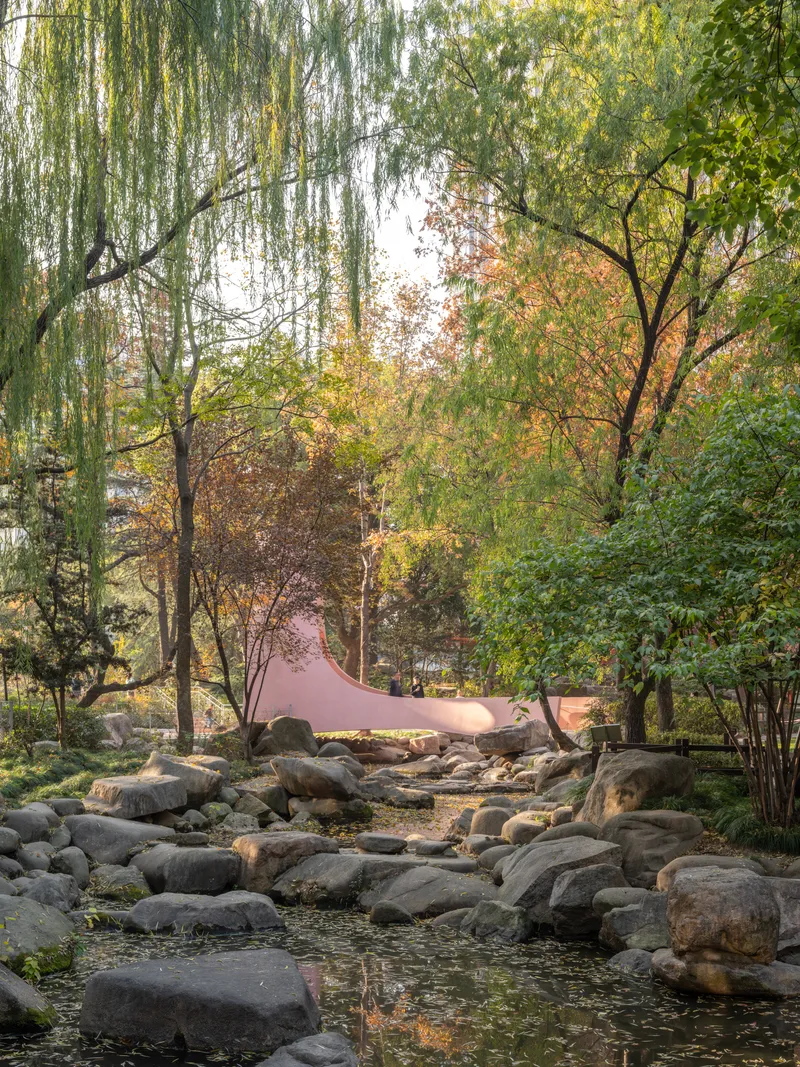 Distant view of the pink structure through willow trees and rocky streambed in soft light