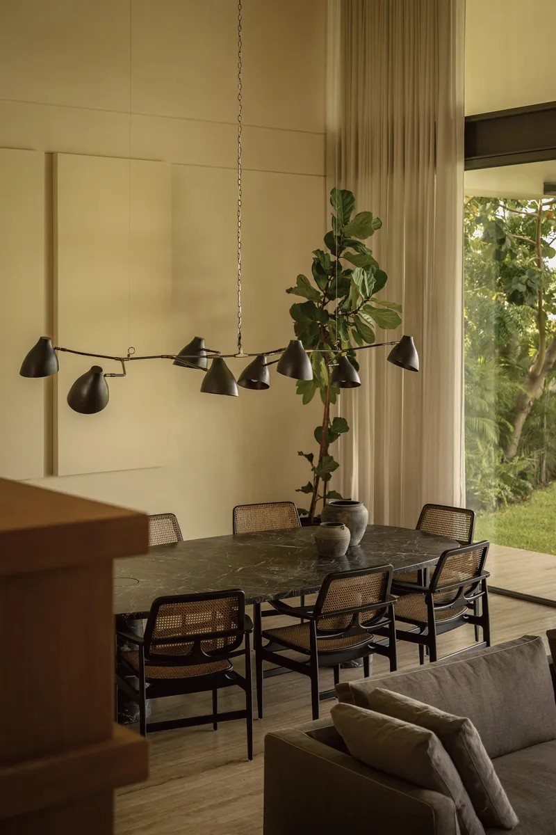 Dining area with chain-suspended chandelier above dark table overlooking garden through sheer curtains