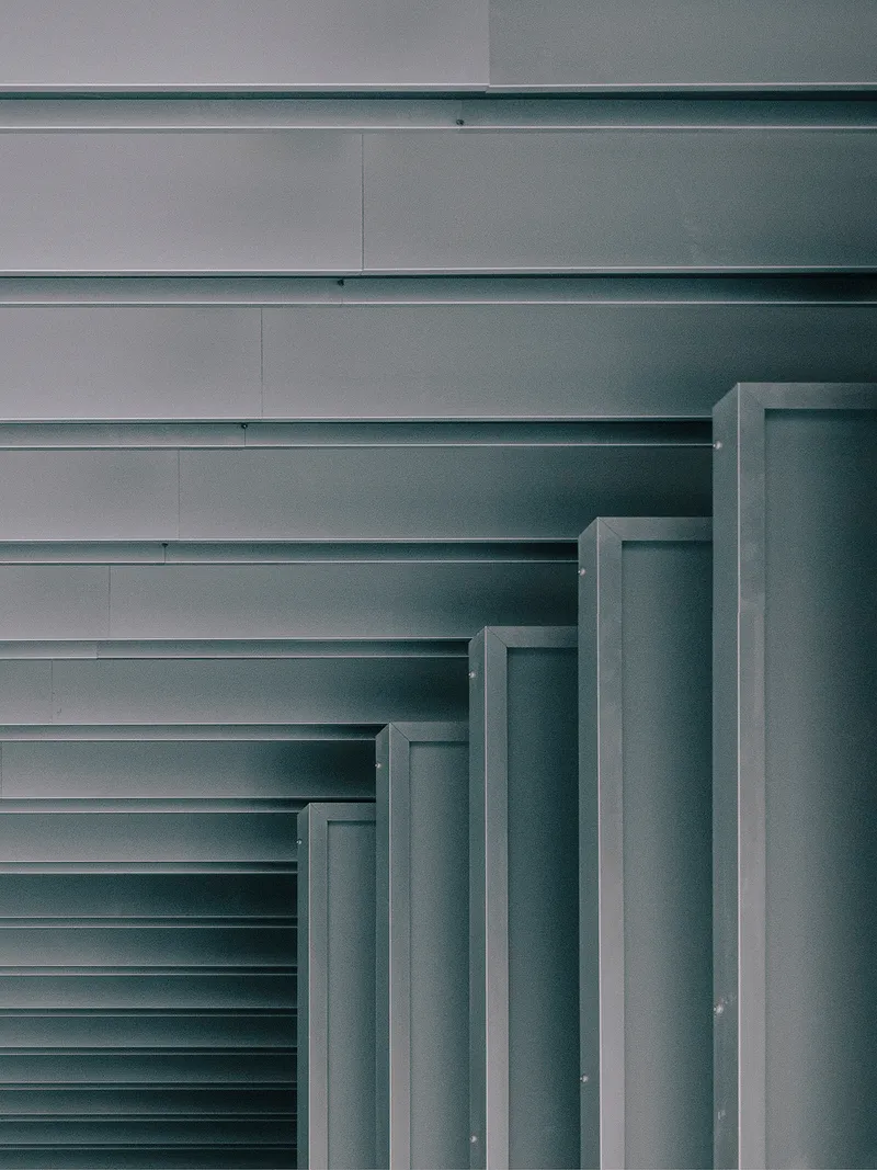 Close-up of stepped metal louvers ascending against horizontal ribbed metal cladding in shadow