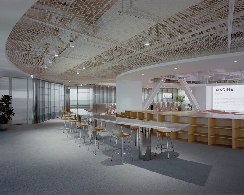 Communal workspace with long tables and white structural steel trusses beneath woven ceiling panels