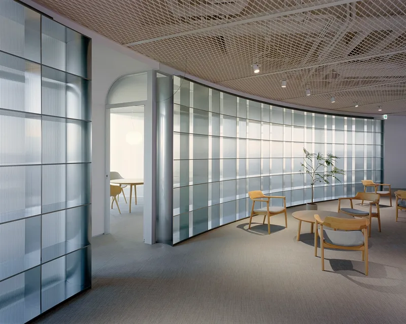 Interior lounge with curved translucent glass panel wall and expanded metal ceiling under recessed lighting
