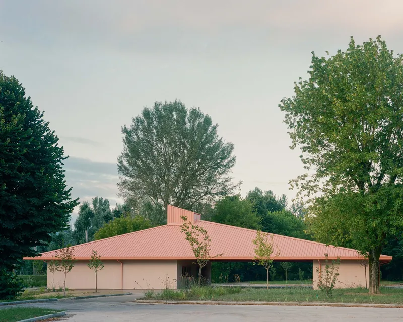 Single-storey structure with pink corrugated metal roof and stucco walls framed by tall deciduous trees