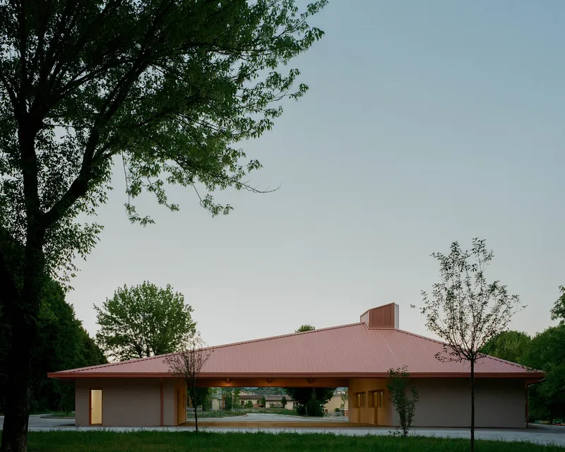 Pavilion with broad hipped metal roof and open-air loggia framed by young trees at dusk