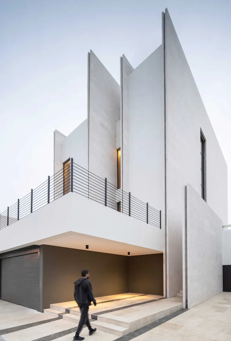 White concrete facade with jagged peaks and a person walking through the covered garage entry at dusk