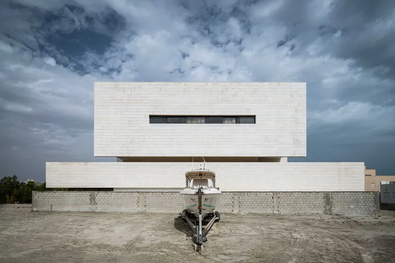 Layered white limestone facade with horizontal slot window and boat centered in forecourt under stormy clouds