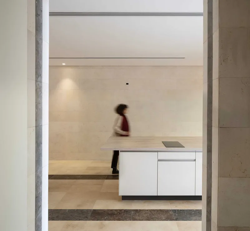 View through doorway of figure in motion crossing pale stone flooring past white kitchen cabinetry