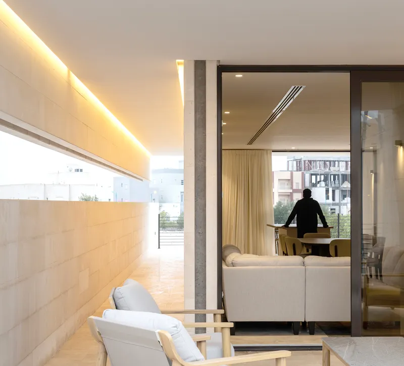 Covered terrace with concealed lighting looking through glazed doors to living room with seated figure