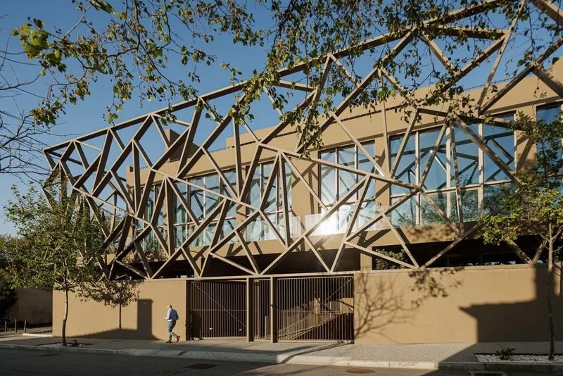 Street view of the diagrid steel frame rising above a tan perimeter wall with trees overhead